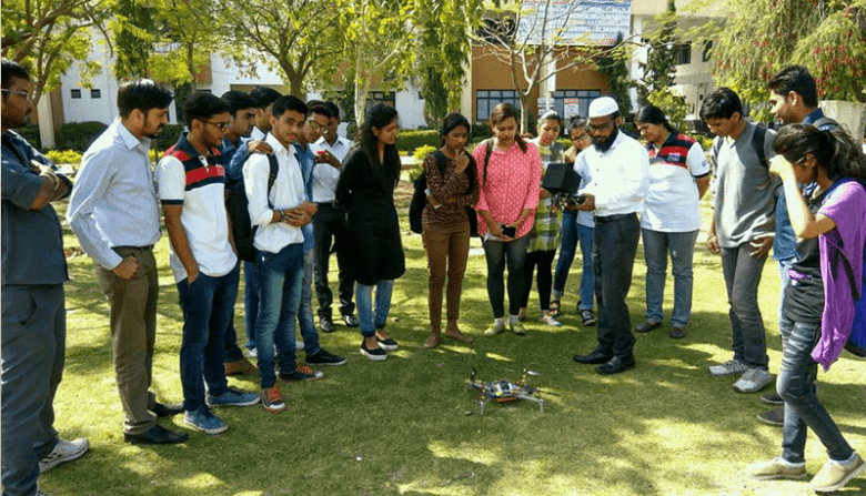 Unmanned technology | tycoonstory media Unmanned technology group of students gathered outdoors, watching a professor demonstrate a drone in action, symbolizing hands-on learning in technology and innovation.