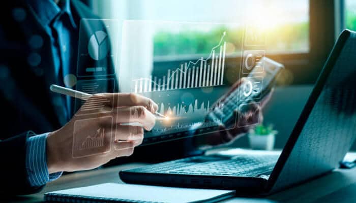 S | tycoonstory media A professional analyzing digital charts and graphs on a transparent screen while working on a laptop, illustrating a detailed financial forecast.
