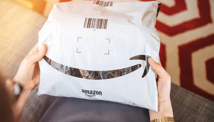 Amazon A person holding a white shipping pouch with the amazon smile logo and a printed barcode on the top, sitting indoors with part of a patterned rug visible in the background.