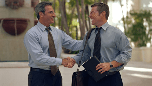 Long term jobs - tycoonstory | tycoonstory media Two businessmen smiling and shaking hands outside an office, symbolizing reduced workplace stress through positive professional relationships.