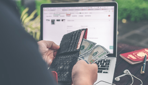Online sales vat-tycoonstory | tycoonstory media Person taking cash from a wallet in front of a laptop. Managing finances and vat payments.