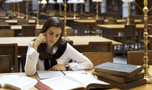 Hire wrting services online -tycoonstory | tycoonstory media A female student, dressed in a white shirt and dark vest, sits at a large wooden table in a dimly lit library, studying open books and papers with her hand resting on her face.