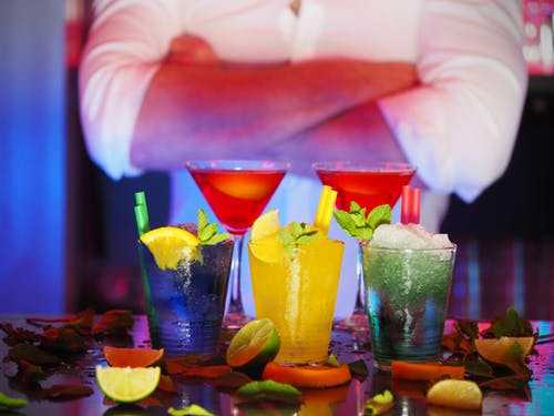 Task Colorful cocktails arranged on a bar counter with a bartender in the background, representing travel jobs in hospitality.