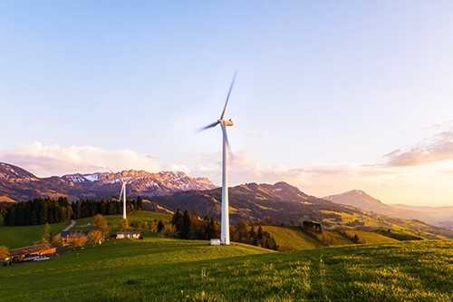 Renewable energy -tycoonstory | tycoonstory media Wind turbines spinning on a green landscape with mountains in the background during sunset, representing renewable energy.