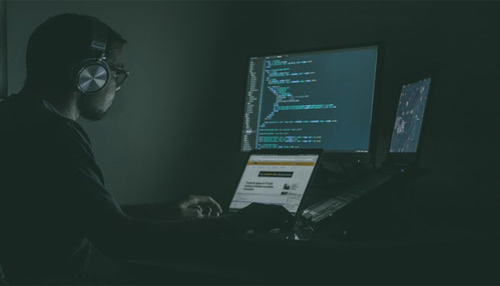 It experts A developer wearing headphones working at multiple monitors in a dimly lit room, writing and debugging code.