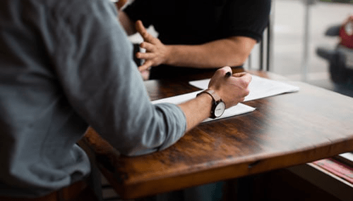 Partners - tycoonstory | tycoonstory media Two entrepreneurs signing documents at a wooden table while discussing a business agreement.