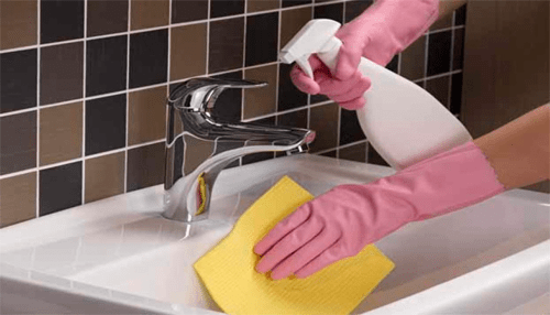 Make cleaning a regular routine Hands wearing gloves cleaning a bathroom sink with spray cleaner and yellow cloth.