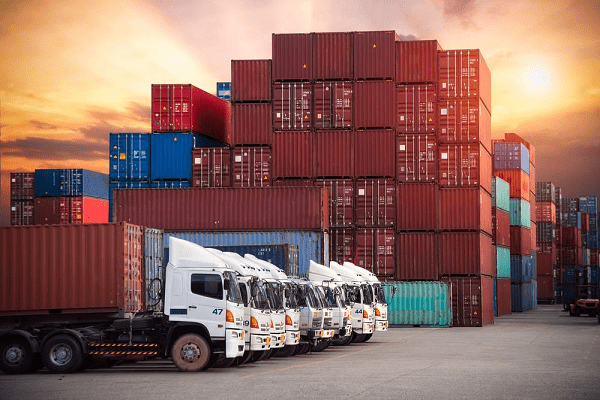 Trucking-services | tycoonstory media Fleet of trucks parked in front of stacked shipping containers at a heavy haul trucking company site.