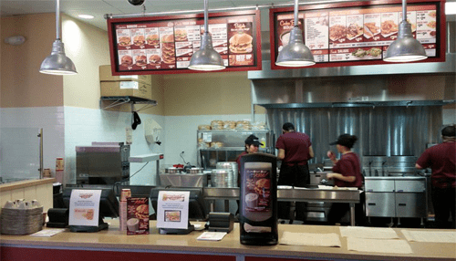 Reduce costs - tycoonstory | tycoonstory media Fast-food counter featuring digital signage menu displays showing meal items while staff prepare food in the kitchen.