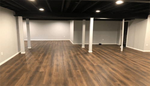 Small designs - tycoonstory | tycoonstory media Spacious basement renovation featuring new dark wood-look flooring, freshly painted light gray walls, recessed lighting, and exposed black ceiling beams supported by white columns