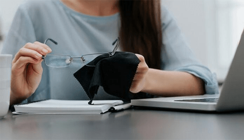 The highs and lows of wearing prescription glasses Person cleaning their prescription glasses while working on a laptop, representing the regular maintenance required for glasses.