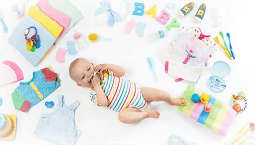 How to financially plan for a new baby A happy baby playing surrounded by colorful baby clothes and accessories, illustrating the variety of essential items parents must financially plan for when welcoming a new child.