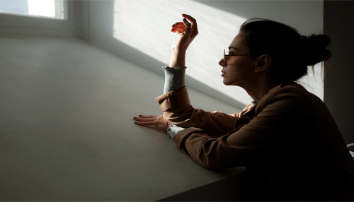 How to return to work while grieving the death of a parent Woman wearing glasses looks out a window, bathed in sunlight, contemplating her return to work.