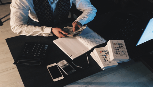 Fractional cfo A fractional cfo reviewing financial records at a desk, counting cash beside a notebook, calculator, laptop, and several smartphone boxes