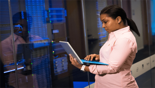 Business intelligence programmer - tycoonstory | tycoonstory media A woman in a pink shirt stands in a server room, holding and working on a slim tablet computer with a blue keyboard, with her reflection visible in the glass panels of the servers behind her.