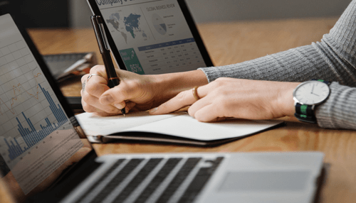 Close-up of a person writing in a notebook with a pen, surrounded by laptops displaying data and graphs on a wooden table, indicating a professional work environment The importance of bookkeeping for a successful property manager