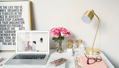 Stylish desk setup featuring a laptop, pink roses in a vase, a gold lamp, and office supplies, creating a vibrant workspace Modern home office desk setup with laptop, flowers, lamp, notebooks, and stylish workspace décor.