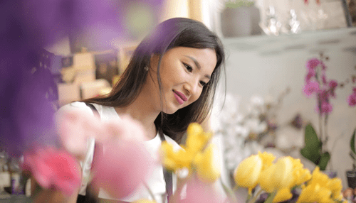 Sourcing the suppliers - tycoonstory | tycoonstory media A woman smiling while arranging colorful flowers, representing product sourcing and global supply opportunities in the import/export business.