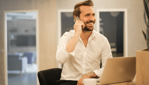 Ask for help - tycoonstory | tycoonstory media Smiling man in a white shirt on a phone call, using a laptop. Blurring the lines between work and private life.