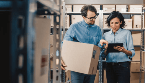 Inventory management Two warehouse employees discussing stock details while checking a tablet and carrying a cardboard box.