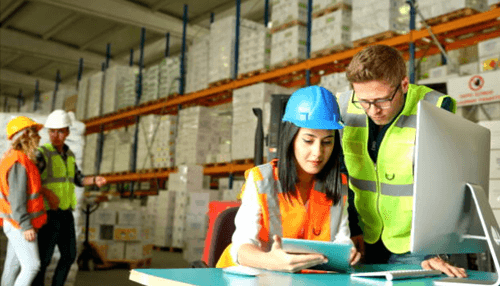 Perpetual inventory management system - tycoonstory | tycoonstory media Workers in safety gear reviewing data on a computer inside a warehouse with shelves stacked with inventory.