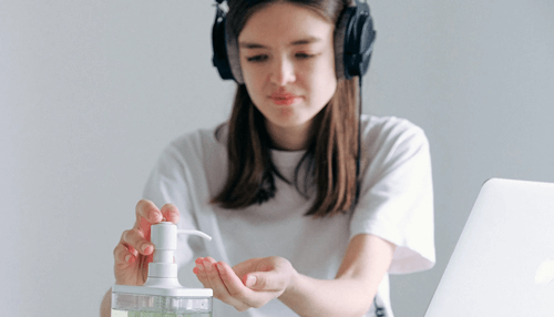 Work from home on virus -tycoonstory | tycoonstory media A woman wearing headphones using hand sanitizer while working on a laptop connected to the internet.