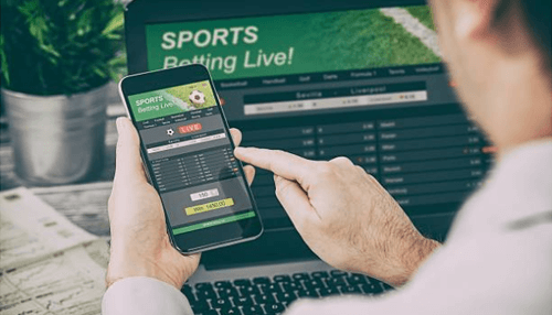 Major companies in betting - tycoonstory | tycoonstory media A close-up of a person holding a smartphone and using an online betting application to view live sports odds, with a laptop screen in the background displaying additional game statistics.