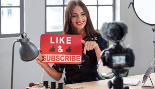 Social media marketing Social media marketing content creator holding a “like & subscribe” sign while recording a video with a camera setup in a modern workspace.
