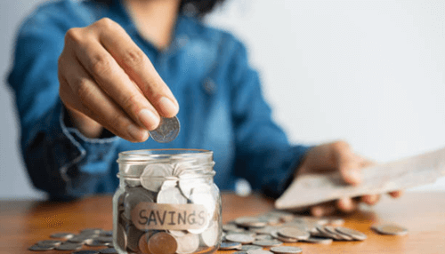Minimal investment A person dropping a coin into a glass jar labeled "savings," illustrating the concept of wealth building starting with a minimal investment.