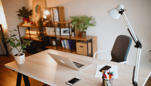 Computer table-tycoonstory | tycoonstory media A bright, well-organized home office setup featuring a light-colored wooden desk with a modern laptop, a smartphone, a cup of pens, and a height-adjustable task lamp. Shelving in the background holds books and plants, creating a comfortable workspace.