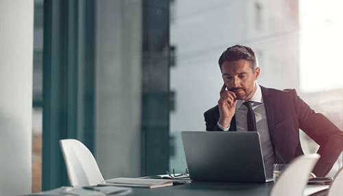 Emotional intelligence A businessman sitting at a desk, talking on the phone while working on a laptop in a modern office setting.