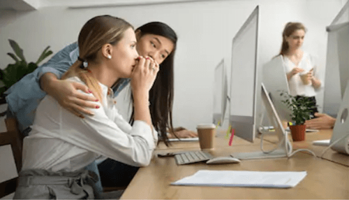 Emotional intelligence Two female coworkers whispering to each other at their desks while another colleague works in the background.