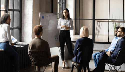 Mistakes HR Managers A business professional presenting ideas to a team during a meeting, standing near a whiteboard while colleagues listen attentively in a modern office setting.