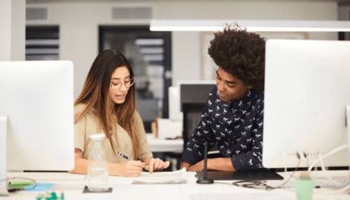 One on one meeting Two colleagues collaborating at a desk in an open office, reviewing notes and discussing work tasks together.