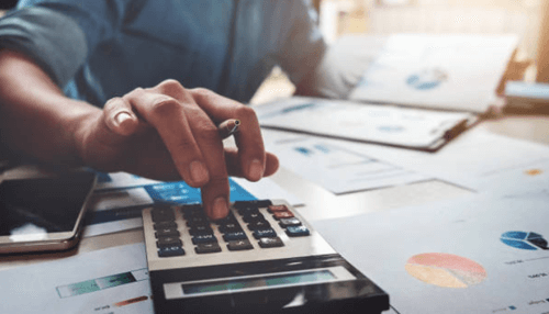 Cpa help startup business-tycoonstory | tycoonstory media A close-up of a hand working on a calculator, surrounded by financial charts and graphs, highlighting the auditing process in action.