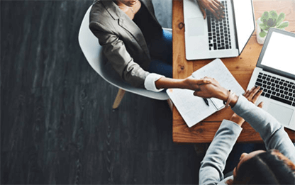 Management experts Two business professionals shaking hands across a desk with laptops and documents, representing merging firms and collaborative partnership.