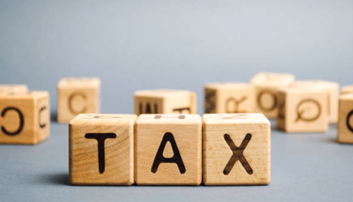 Tax benefits A professional woman with curly hair and glasses wearing a blue blazer stands with her arms crossed, looking thoughtfully out of a high-rise office window overlooking a blurred cityscape.