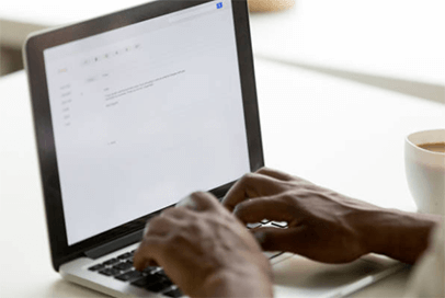 Top essential prompts for business communication-tycoonstory | tycoonstory media A young, white man wearing glasses and a red sweater is sitting at a wooden table in front of a large window, intently working on a laptop. He appears to be focused on writing or reviewing business correspondence in a cafe or office setting.