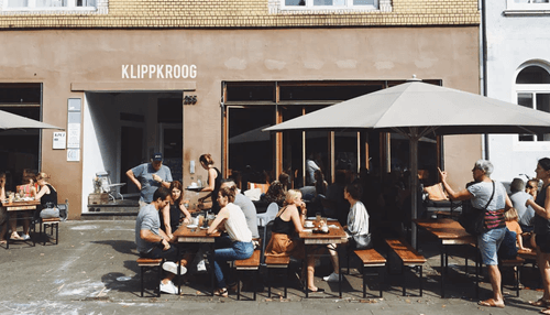 Choose the right location - tycoonstory | tycoonstory media Outdoor restaurant seating with umbrellas and patrons enjoying their meals at long wooden tables, located in a lively urban setting