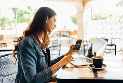 Research before investing in crypto-tycoonstory | tycoonstory media A woman using a smartphone and laptop in a cafe, likely managing bitcoin investments or tracking cryptocurrency trends