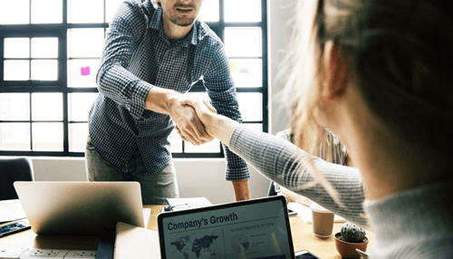 B2b payment solution - tycoonstory | tycoonstory media Two professionals shaking hands across a desk with a laptop displaying charts, symbolizing a b2b payment agreement or business transaction between companies.