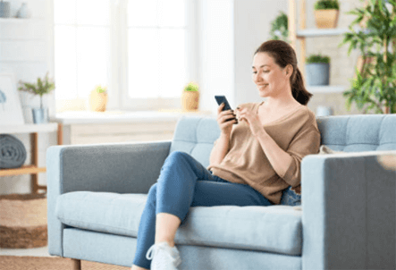 Ecommerce business A smiling woman sitting comfortably on a light blue couch in a bright living room, looking at and interacting with her smartphone.