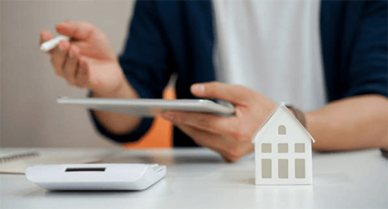 Choosing moving estimate company-tycoonstory | tycoonstory media A person holding a tablet while reviewing moving estimates, with a small house model on the desk, preparing for a move and comparing options.