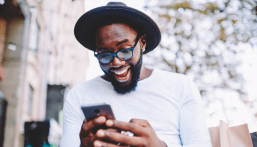 Website is mobile friendly - tycoonstory | tycoonstory media A cheerful man wearing glasses and a hat smiling excitedly while looking at his smartphone outdoors.