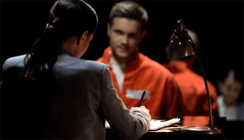 Criminal specialists-tycoonstory | tycoonstory media A criminal defence attorney interviewing a man in a red uniform at a desk with a focused lamp in a dimly-lit room.