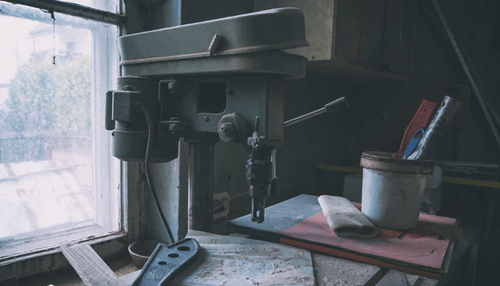 Drilled machines are glued to a bench - tycoonstory | tycoonstory media Old workshop drill press equipment near a window, used for metalworking and precision machining tasks.