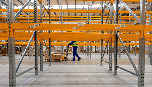 Distribution centers Warehouse worker moving goods with a hand pallet truck beneath the empty, metal shelving structure of a logistics facility.