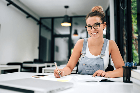 Become a student entrepreneur-tycoonstory | tycoonstory media Smiling student entrepreneur working at a desk, writing in a notebook with a laptop in front of her, reflecting the focused and determined nature of young business founders.