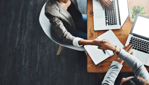 Growing their business - tycoonstory | tycoonstory media A smiling businessman in a grey suit jacket shaking hands with another professional in a modern office, representing a successful partnership or the finalization of a business agreement.