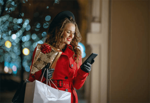 Personalized discount coupons-tycoonstory | tycoonstory media A woman in a red coat smiling while reading a push notification on her phone, with holiday shopping bags in hand, standing in front of festive lights.